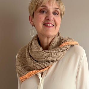 May include: A tan knitted cowl with an orange border is worn around the neck. The cowl is made of a soft, textured yarn. The woman is wearing a white blouse and silver earrings. The background is a solid, neutral color.