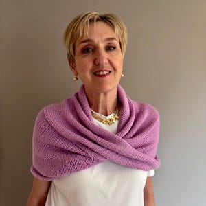 Peut inclure: Une femme portant un châle tricoté violet clair sur un t-shirt blanc. Le châle est drapé autour des épaules et de la poitrine. Elle porte également un collier et des boucles d'oreilles dorés. L'arrière-plan est gris uni.