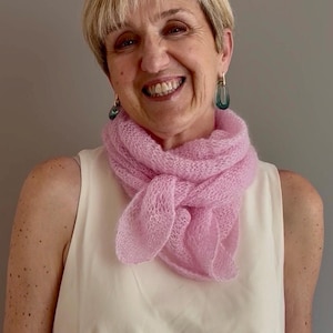 Peut inclure: Un foulard triangulaire tricoté en rose clair, semi-transparent, avec une texture lâche et aérée. Le foulard est enroulé autour du cou et noué simplement sur le devant. La matière semble douce et duveteuse, avec un motif à mailles ouvertes.