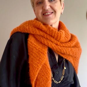 Può includere: Una sciarpa lavorata a maglia di colore arancione vivace e soffice, drappeggiata sulle spalle. La sciarpa presenta una trama larga e testurizzata ed è indossata sopra una camicia nera abbottonata con vita arricciata e una catena dorata.