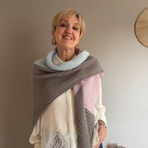 Peut inclure: Une femme porte un châle tricoté gris, bleu clair et rose. Elle porte une blouse blanche avec des détails en dentelle et un jean bleu. Elle porte également des boucles d'oreilles dorées.