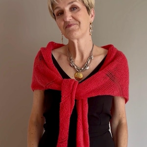 Peut inclure: Un châle tricoté rouge vif drapé sur les épaules et noué sur le devant, porté sur un haut noir sans manches. Le châle présente un motif en maille texturée et lâche avec de longues extrémités tombantes, accompagné d'un collier argenté superposé.