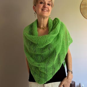 Może przedstawiać: Żywy, zielony szal z dzianiny, narzucony na ramiona. Szal ma teksturowany, ażurowy wzór. Kobieta ma na sobie czarny top i białe spodnie. Ma również złotą biżuterię.