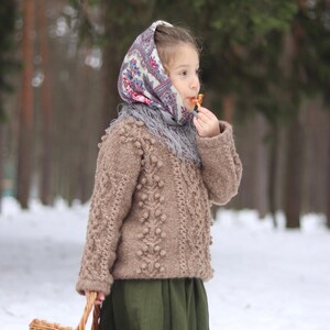May include: A young girl wearing a brown sweater with a patterned headscarf and a green skirt holds a wicker basket in a snowy forest.