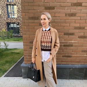 May include: A woman wearing a brown coat, a white shirt, a brown and white checkered sweater, and plaid pants. She is carrying a black handbag and wearing black ankle boots.