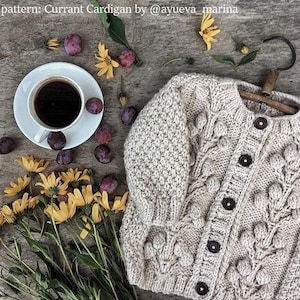 May include: A beige knitted cardigan with a cable knit pattern and large buttons. The cardigan has a textured surface and is decorated with small, round, knitted embellishments. The cardigan is laid out on a wooden surface with a cup of coffee, plums, and yellow flowers.