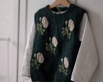 May include: A dark green knitted vest with white embroidered roses. The vest is hanging on a wooden hanger against a white background.
