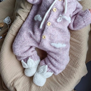 Op de afbeelding: Een roze gebreide baby romper met witte accenten en konijnenoren. De romper heeft een capuchon en knopen aan de voorkant. De voeten van de romper zijn gevormd als konijnenpootjes.