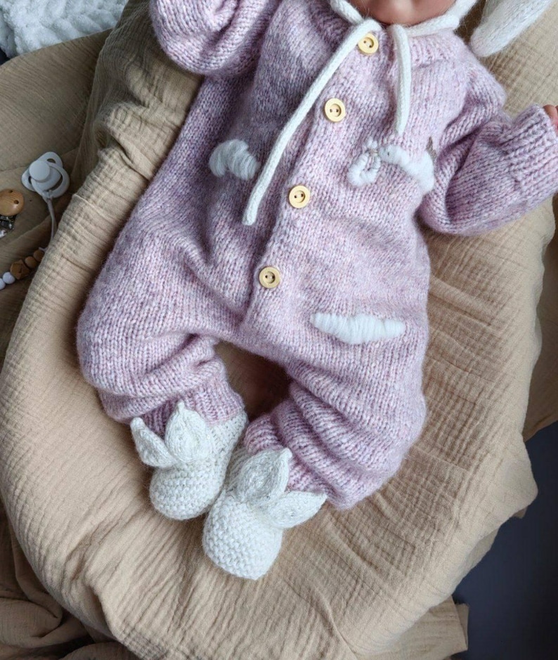 Op de afbeelding: Een roze gebreide baby romper met witte accenten en konijnenoren. De romper heeft een capuchon en knopen aan de voorkant. De voeten van de romper zijn gevormd als konijnenpootjes.