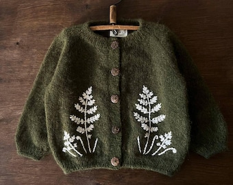 May include: A green knitted cardigan with white embroidered fern designs. The cardigan has a button closure.