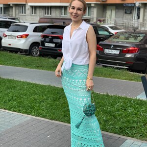 May include: A woman wearing a white sleeveless shirt tied at the waist, a light blue crochet maxi skirt, and a small green and blue purse with a tassel. She is standing on a sidewalk in front of a building.