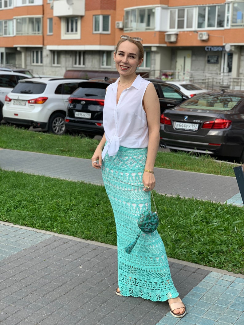 May include: A woman wearing a white sleeveless shirt tied at the waist, a light blue crochet maxi skirt, and a small green and blue purse with a tassel. She is standing on a sidewalk in front of a building.