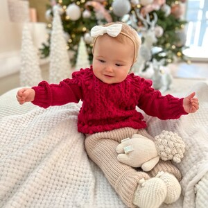 May include: A baby wearing a red knitted sweater with white pom-poms, beige knitted pants, and a white knitted lamb toy. The baby is sitting on a white fluffy blanket in front of a Christmas tree.