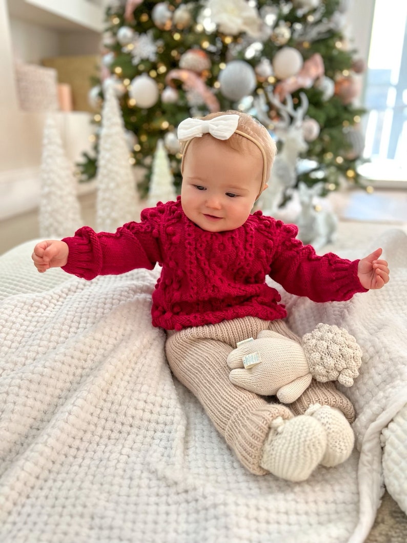 May include: A baby wearing a red knitted sweater with white pom-poms, beige knitted pants, and a white knitted lamb toy. The baby is sitting on a white fluffy blanket in front of a Christmas tree.