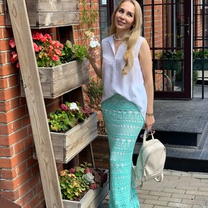May include: A woman wearing a white sleeveless shirt and a light blue crochet maxi skirt. She is standing in front of a brick wall with a wooden plant stand.
