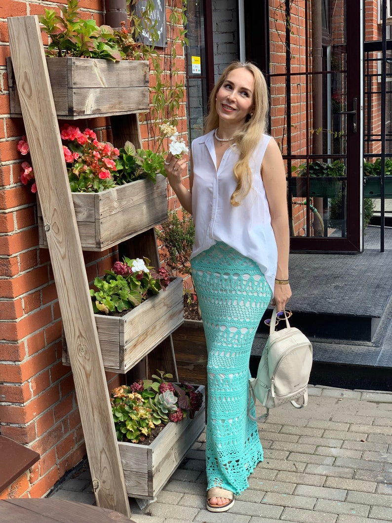 May include: A woman wearing a white sleeveless shirt and a light blue crochet maxi skirt. She is standing in front of a brick wall with a wooden plant stand.