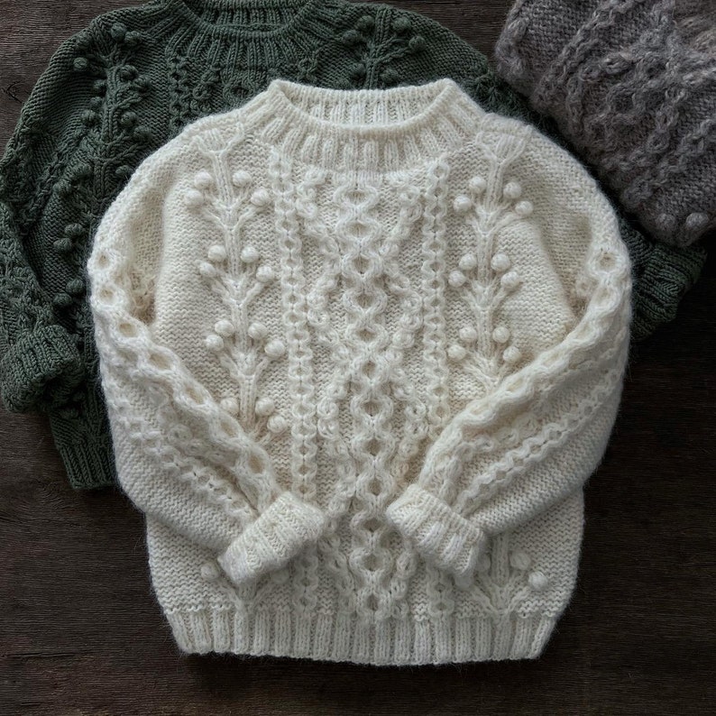 May include: A white knitted sweater with a cable knit pattern and small white pom-poms. The sweater has a crew neck and long sleeves.