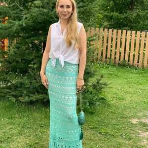 May include: A woman wearing a white sleeveless shirt tied at the waist and a long, crocheted, turquoise skirt. She is standing in a grassy area with a wooden fence in the background.