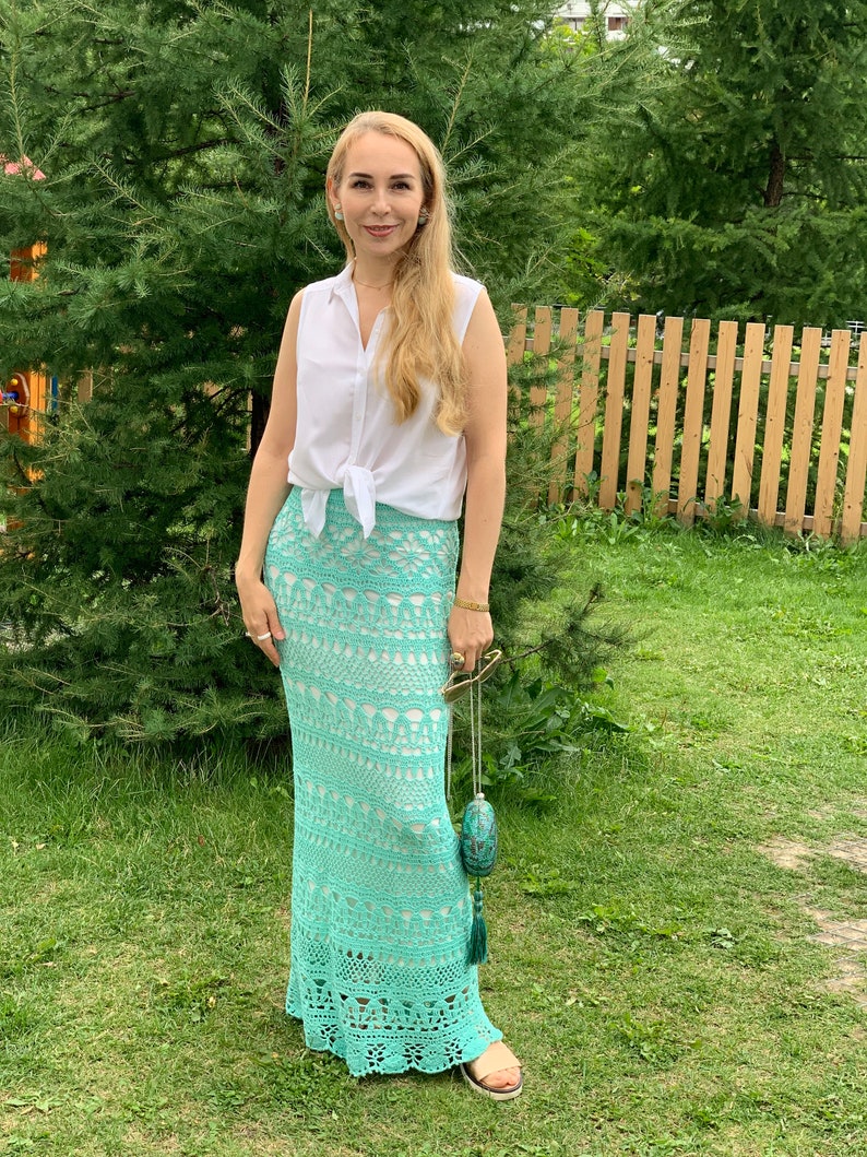 May include: A woman wearing a white sleeveless shirt tied at the waist and a long, crocheted, turquoise skirt. She is standing in a grassy area with a wooden fence in the background.