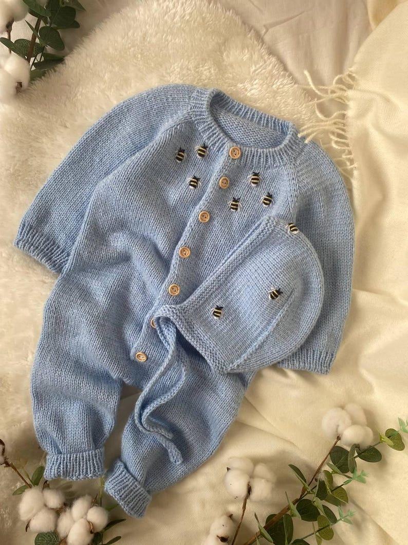 Op de afbeelding: Een lichtblauwe gebreide baby-outfit met een bijpassende muts. De outfit heeft een knoopsluiting en is versierd met geborduurde bijen.