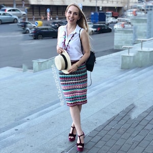 May include: A woman wearing a white sleeveless shirt, a colorful striped skirt, and burgundy heeled sandals. She is holding a straw hat and a black backpack. The woman is standing on a set of stairs.