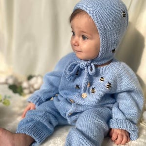 Op de afbeelding: Een baby draagt een lichtblauwe gebreide outfit met een bijpassende muts. De outfit en de muts zijn versierd met geborduurde bijen. De baby zit op een wit, pluche oppervlak.