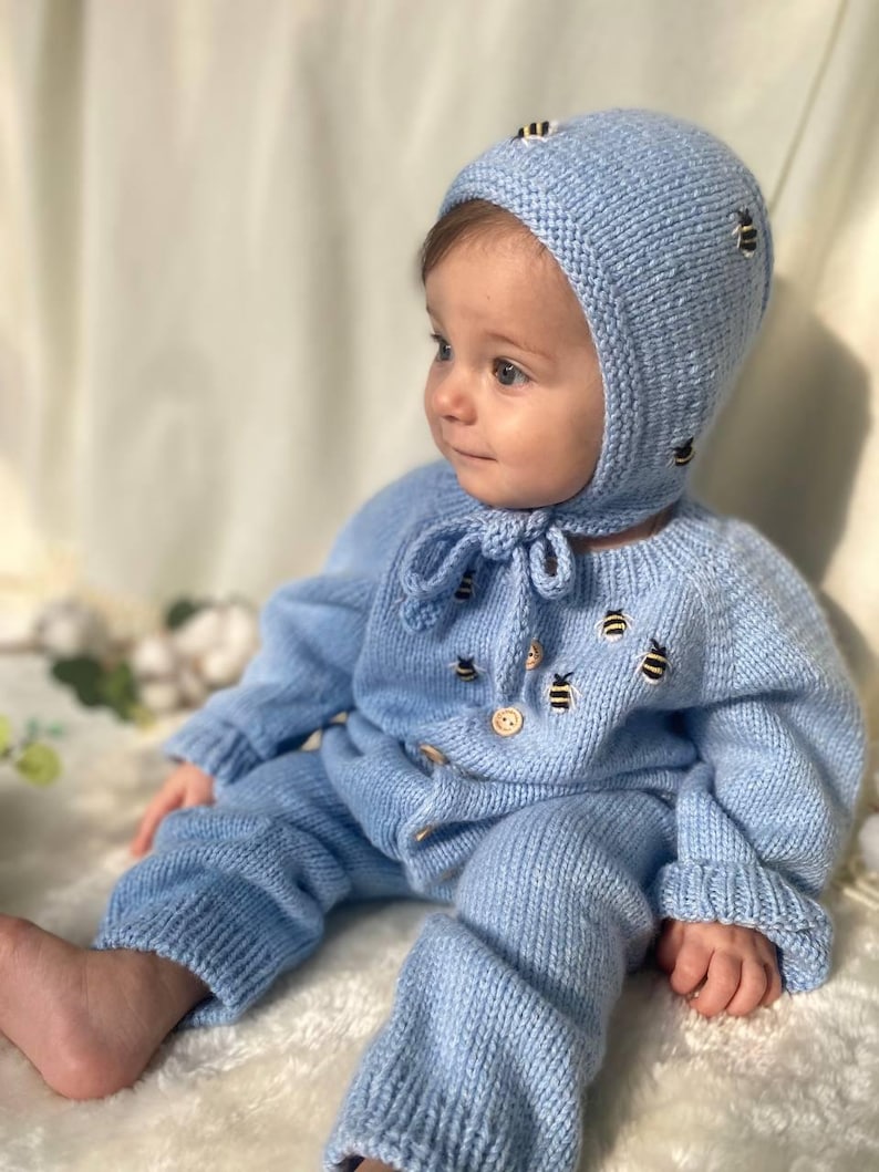 Op de afbeelding: Een baby draagt een lichtblauwe gebreide outfit met een bijpassende muts. De outfit en de muts zijn versierd met geborduurde bijen. De baby zit op een wit, pluche oppervlak.
