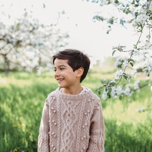 May include: A young boy wearing a light brown cable knit sweater with white pom-poms, smiling and looking to the right. He is standing in a field of green grass with white flowers in the background.