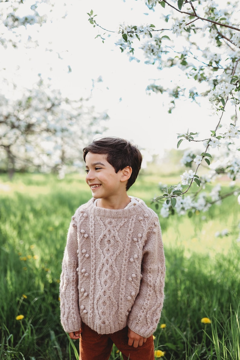 May include: A young boy wearing a light brown cable knit sweater with white pom-poms, smiling and looking to the right. He is standing in a field of green grass with white flowers in the background.