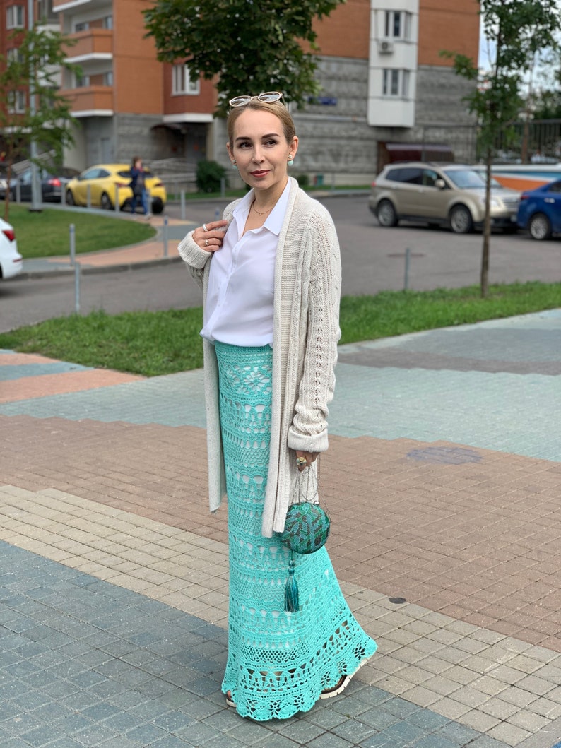 May include: A woman wearing a white button-down shirt, a beige cardigan, and a long, teal crochet skirt. She is holding a teal and green beaded clutch.
