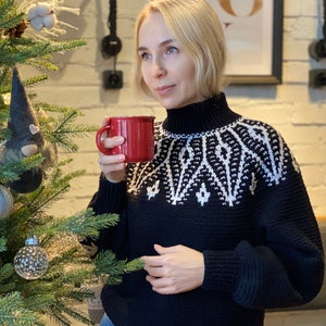 May include: A woman wearing a black sweater with a white geometric pattern around the neck. She is holding a red mug and looking to the right. A Christmas tree is in the background.