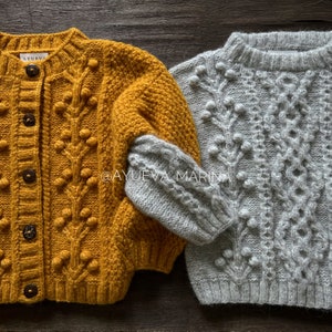 May include: Two knitted jumpers with a cable knit pattern and pom pom accents. One jumper is mustard yellow and the other is light grey. The jumpers are laid out on a wooden surface. The text "@ayueva_marina" is visible on the image.