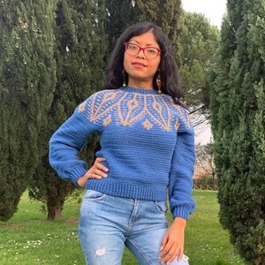 May include: A blue crocheted sweater with a geometric pattern in gold and beige. The sweater has a crew neck and long sleeves.