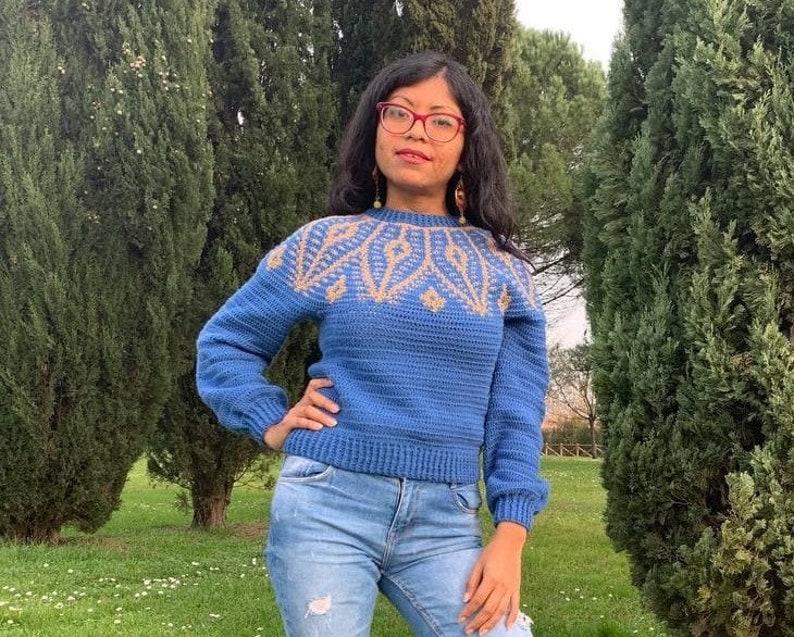May include: A blue crocheted sweater with a geometric pattern in gold and beige. The sweater has a crew neck and long sleeves.