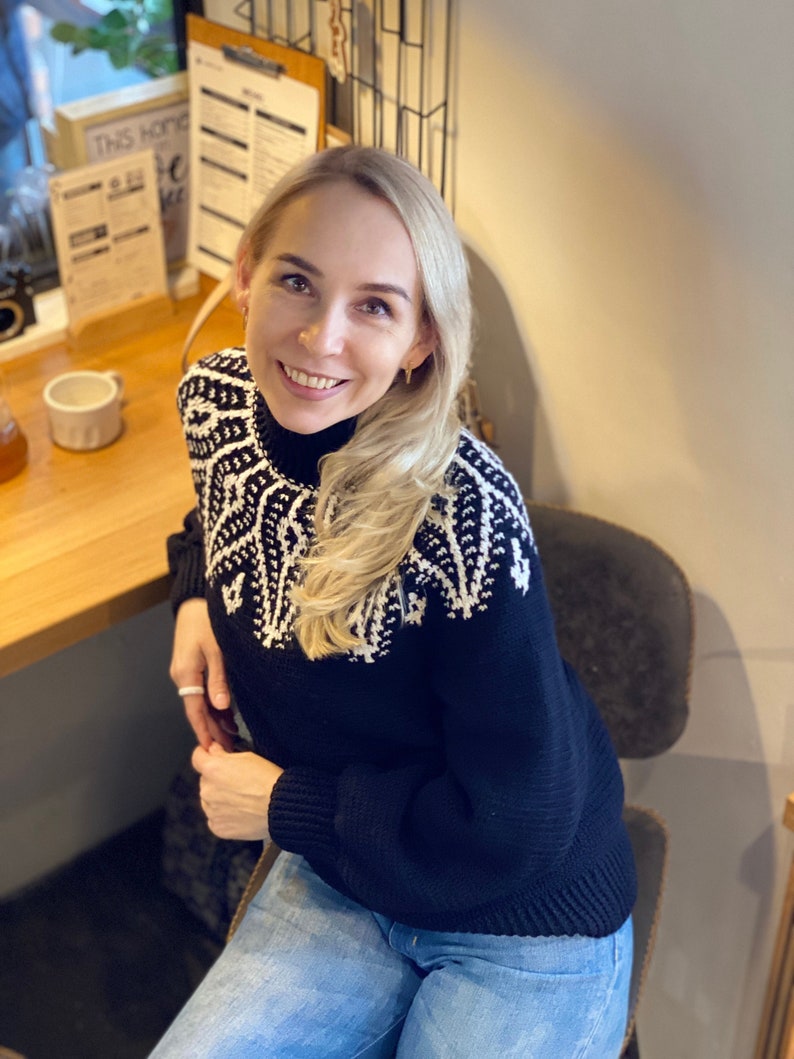 May include: A woman wearing a black and white patterned sweater with a mock neck and blue jeans. She is sitting in a chair and looking at the camera.