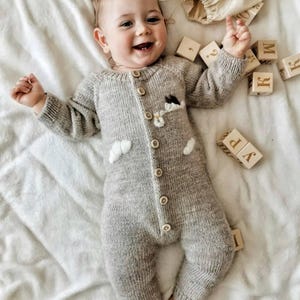 Op de afbeelding: Een baby draagt een grijze gebreide romper met een wolk- en bloemetjesontwerp. De baby ligt op een wit deken en speelt met houten blokken die de letters "W", "P", "K" en "R" spellen.