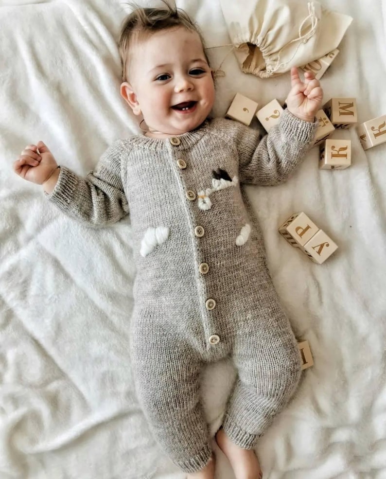 Op de afbeelding: Een baby draagt een grijze gebreide romper met een wolk- en bloemetjesontwerp. De baby ligt op een wit deken en speelt met houten blokken die de letters "W", "P", "K" en "R" spellen.