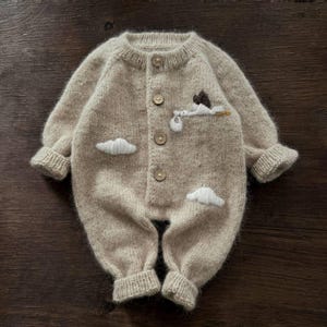 Op de afbeelding: Een beige gebreide baby romper met een witte geborduurde ooievaar die een baby en wolken draagt. De romper heeft een knoopsluiting aan de voorkant.