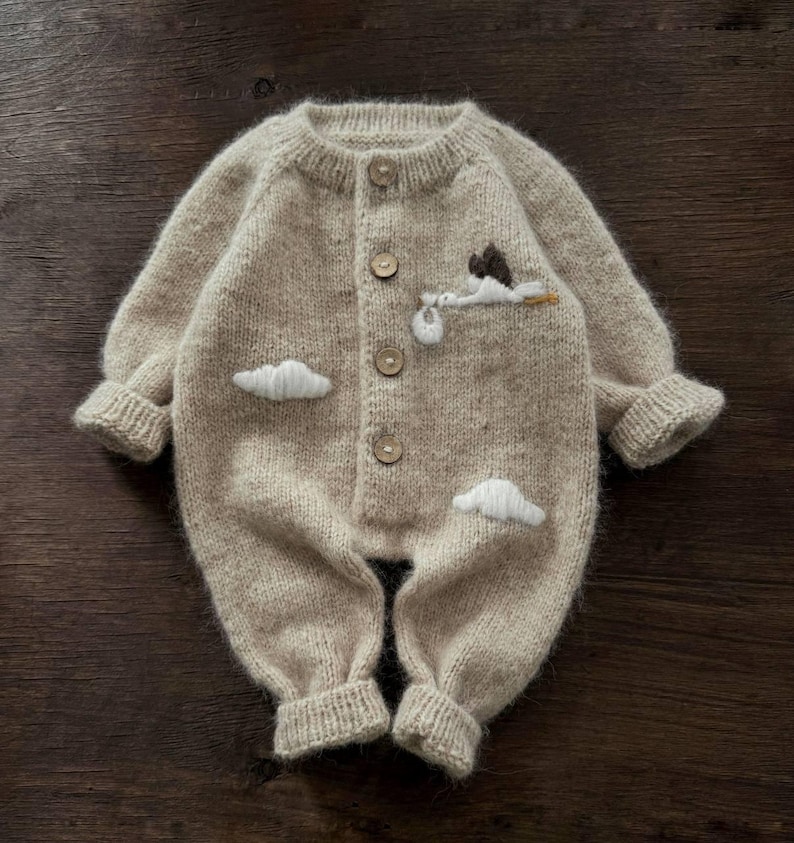 Op de afbeelding: Een beige gebreide baby romper met een witte geborduurde ooievaar die een baby en wolken draagt. De romper heeft een knoopsluiting aan de voorkant.