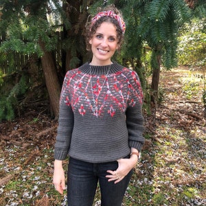 May include: A woman wearing a gray sweater with a red and white geometric pattern, black jeans, and a red and white headband. She is standing in front of a green bush.