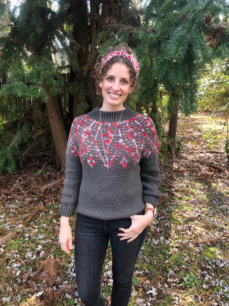 May include: A woman wearing a gray sweater with a red and white geometric pattern, black jeans, and a red and white headband. She is standing in front of a green bush.