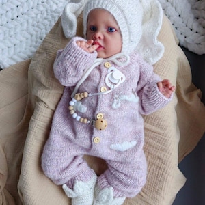 Op de afbeelding: Een baby draagt een roze gebreide bunny-thema onesie met witte accenten en een witte gebreide bunny-hoed. De baby houdt een speen vast en heeft een houten kralenketting.