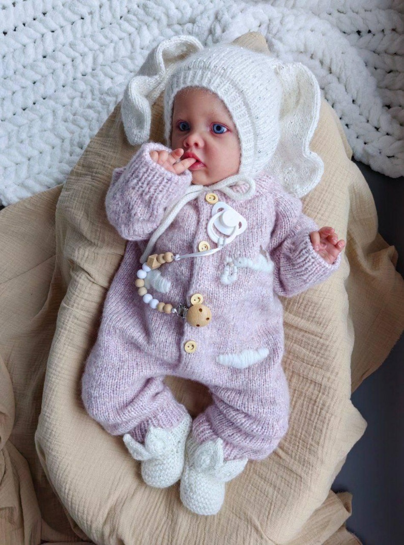 Op de afbeelding: Een baby draagt een roze gebreide bunny-thema onesie met witte accenten en een witte gebreide bunny-hoed. De baby houdt een speen vast en heeft een houten kralenketting.
