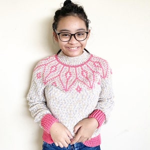 May include: A young person wearing a pink and white knitted sweater with a geometric pattern. The sweater has pink trim on the cuffs and neckline.