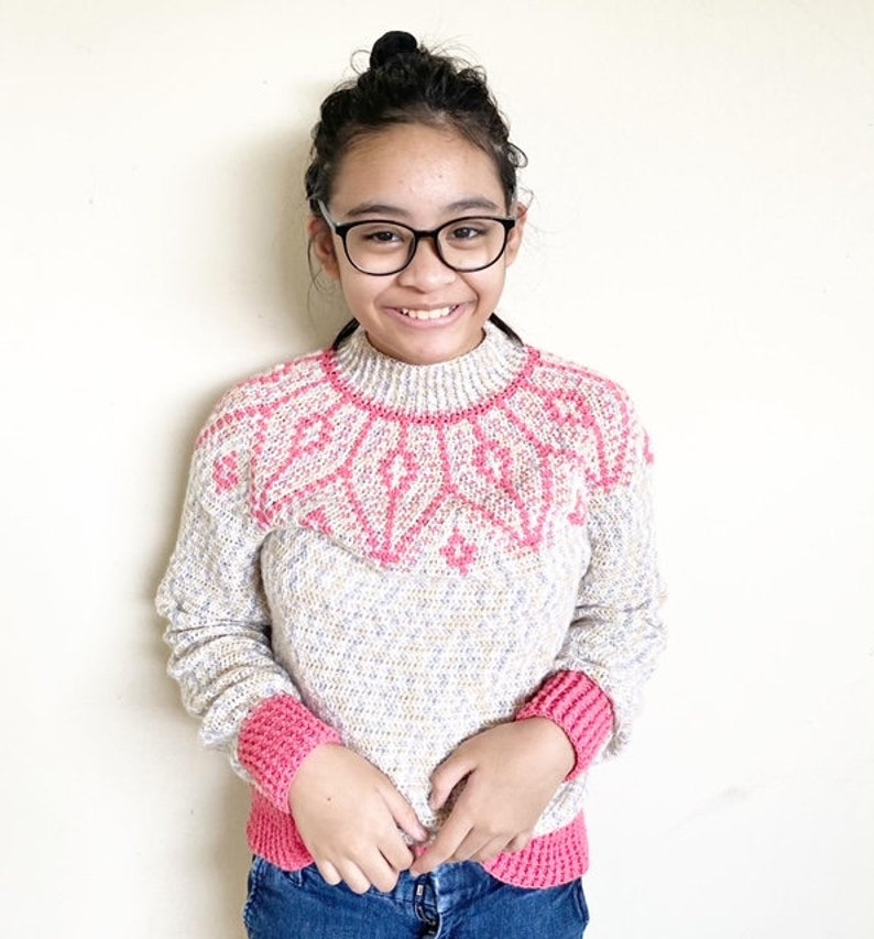 May include: A young person wearing a pink and white knitted sweater with a geometric pattern. The sweater has pink trim on the cuffs and neckline.