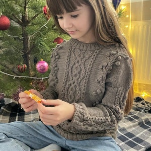 May include: A young person wearing a brown knitted sweater with a cable knit pattern and pom poms. They are sitting on a plaid blanket in front of a Christmas tree.