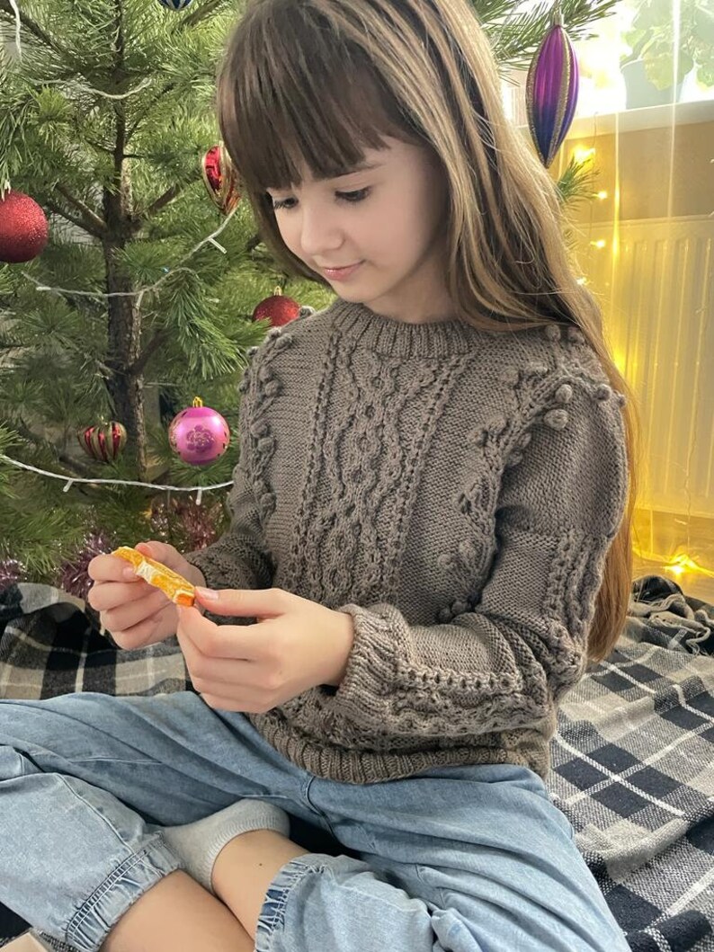 May include: A young person wearing a brown knitted sweater with a cable knit pattern and pom poms. They are sitting on a plaid blanket in front of a Christmas tree.