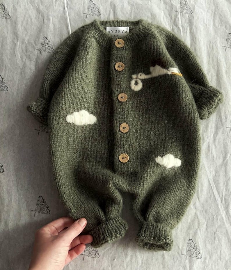 Op de afbeelding: Een groene gebreide baby romper met witte wolk en vogel applicaties en houten knopen. De romper heeft de tekst "AYUEVA" op de rug.