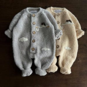 Könnte beinhalten: Zwei gestrickte Baby-Strampler, einer grau und einer beige, mit gestickten Wolken und Vögeln. Die Strampler haben Knöpfe zum Verschließen an der Vorderseite.
