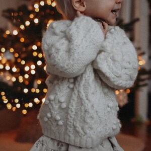 May include: A young child wearing a white knitted sweater with white pom-poms and a white and gray floral skirt. The child is standing in front of a Christmas tree with lights.